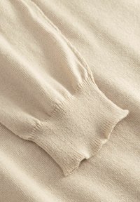 Gros plan sur la manche d'un pull en tricot de couleur crème avec un poignet côtelé et des détails subtils de couture verticale.