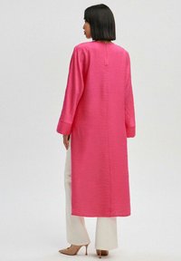 Largo vestido rosa con un acabado texturizado, que cuenta con mangas largas y cierre en la parte posterior, combinado con pantalones blancos y tacones nude.