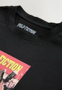 T-shirt noir avec un graphisme de Pulp Fiction et un texte jaune sur le devant, avec le titre du film imprimé à l'intérieur du col.