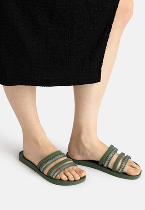 Füße, die grüne Slide-Sandalen mit drei strukturierten Riemen tragen, kombiniert mit einem schwarzen strukturierten Rock vor weißem Hintergrund.