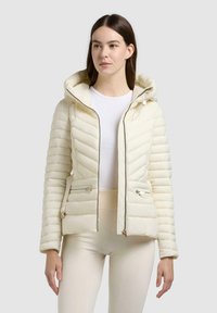 Femme portant une doudoune à capuche écrue sur une chemise blanche et un pantalon couleur crème, debout devant un fond gris uni.