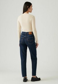Geribbelde crèmekleurige longsleeve top gecombineerd met donkerblauwe high-waisted jeans met een leren patch en Levi's kenmerkende achterzakken. Zwarte schoenen.
