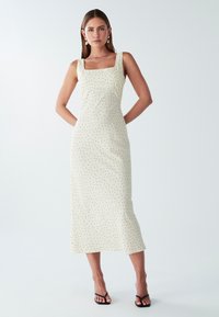 Žena stoji s rukama iza leđa, odjevena u bezrukavnu krem haljinu s crnim točkama i crnim sandalama na visoku petu, prema bijeloj pozadini.