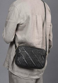 Bolso bandolera negro tejido con forma rectangular, que presenta una superficie texturizada, una correa delgada y acentos en dorado.