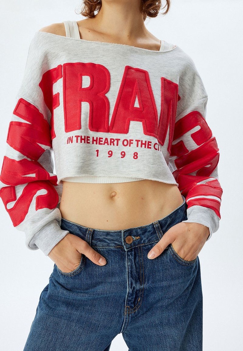 Personne portant un sweat gris court avec de grandes lettres rouges et un jean bleu taille haute, mains dans les poches, ventre apparent.