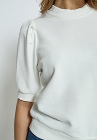 Vit, lätt sweatshirt med hög, ribbad krage, korta puffärmar och ribbat nederkant. Slät textur med minimal detaljer.