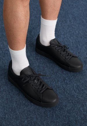 Fekete bőr sportcipők kerek orral és lapos talppal. Fekete fűzők vannak rajtuk, és fehér crew zoknival vannak párosítva egy kék szőnyegen.