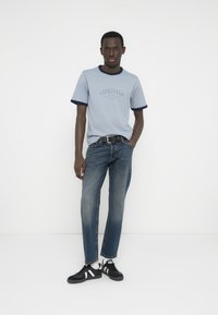 Γαλάζιο μπλουζάκι με σκούρο ναυτικό μπλε περιμετρικό γύρω από τον λαιμό και τα μανίκια, με τη λέξη «ORIGINALS» τυπωμένη στο κέντρο. Φορεμένο με ξεθωριασμένο μπλε τζιν και μαύρα αθλητικά παπούτσια.