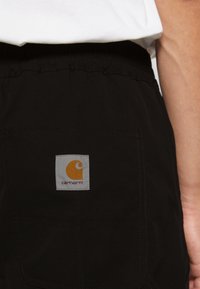 Czarne spodnie z elastycznym paskiem; z tyłu kieszeń z białą kwadratową łatą z pomarańczowym logo. Gładka faktura.