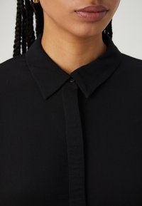 Svart blus med klassisk krage, framknäppning och mjukt, lättviktigt tyg. Detaljer inkluderar subtil textur och inga synliga mönster.