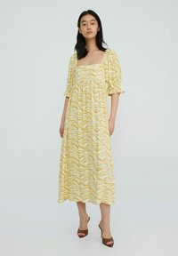 Gele maxi-jurk met een golvend wit patroon, vierkante halslijn, pofmouwen en gathered taille. Model draagt bruine hakken.