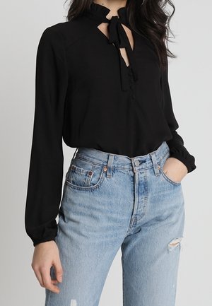 Zwarte blouse met een geknoopte hals, lange mouwen, subtiele ruches en knoopdetails, gecombineerd met lichtblauwe distressed jeans.