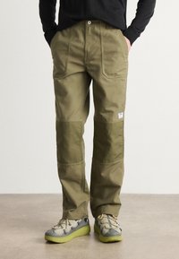Pantalones cargo verde oliva de corte relajado, con grandes bolsillos delanteros y parches en las rodillas de color contrastante. Combinados con zapatillas grises y lima.