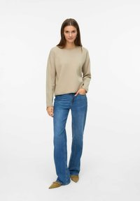 Maglione beige a maniche lunghe abbinato a jeans blu a palazzo e scarpe beige a punta. Design semplice, texture liscia e palette di colori neutri.