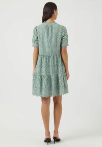 Svetlo zelena obleka s očesi, kratkimi pufastimi rokavi, večplastno zasnovo, valovitim robom in zapletenim perforiranim vzorcem. Prikazan zadnji pogled.