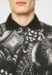 Czarna i biała koszulka polo z wzorem paisley oraz printem zwierzęcym, z kontrastowym czarnym kołnierzykiem i teksturowanym emblematem na piersi.