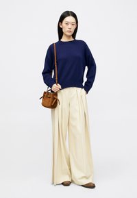Pull en maille bleu marine, pantalon plissé blanc, sac seau en suède marron avec cordon de serrage, et chaussures plates marron. Silhouette simple et décontractée.