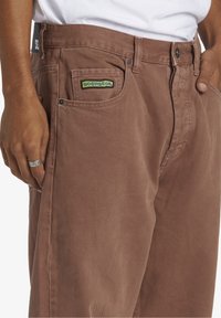 Bruna manchesterbyxor med en lätt glans, med en blå och gul DC Shoe Co-lapp på vänster framficka och standarddetaljer i metall.