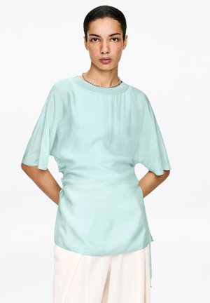 Frau mit dunklem, zurückgebundenem Haar, die eine hellblaue, lockere Bluse und weiße Hose trägt, mit den Händen hinter dem Rücken stehend vor weißem Hintergrund.