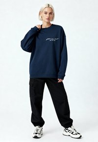Femme blonde portant un sweat-shirt bleu marine avec le texte "suis ton cœur", un pantalon cargo noir et des baskets blanches et noires, debout devant un fond blanc.