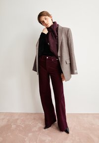 Grijs textuur blazer over een zwarte top, gecombineerd met bordeaux corduroy flared broeken en zwarte spitse hakken. Zachte achtergrond met een roze vloer.
