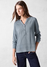 Blusa leggera con bottoni, a righe verticali blu e bianche, con scollatura a V poco profonda e maniche lunghe, abbinata a jeans scuri.