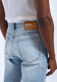 Los jeans de denim azul claro presentan un acabado texturizado, un parche de cuero marrón con la marca y bolsillos traseros estándar con detalles de costura sutiles.