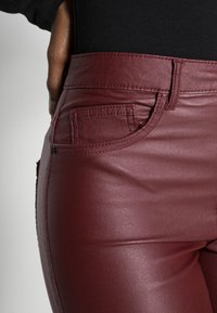 Burgundy umělé kožené kalhoty s přiléhavým střihem, obsahující přední kapsu, jemný lesk a kontrastní prošívání podél švů.