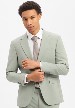 Mann, der den Ärmel eines hellgrauen Anzugs justiert, trägt ein weißes Hemd und eine beige gemusterte Krawatte, vor schlichtem weißem Hintergrund.