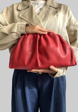 Bolso de cuero rojo con parte superior fruncida, textura suave y forma redondeada, sostenido frente a una persona que lleva una chaqueta beige y jeans azules.