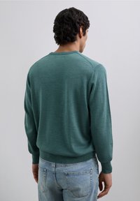 Grön stickad tröja med långa ärmar, rund halsringning samt ribbade muddar och fåll. Bärs med blå jeans i denim som visar en slät textur.