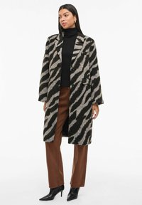 Kvinde iført en lang zebra-stribet frakke, sort turtleneck, brune bukser og sorte spidse hæle, stående med afslappede hænder.