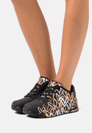 Schwarze Sneaker mit einem gemusterten Design, das weiße und goldene Herzformen zeigt, aus strukturiertem Material und mit schwarzen Schnürsenkeln. Gummisohle für guten Halt.