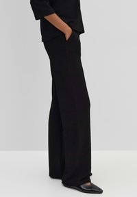 Zwarte wijde pijpenbroek van zachte stof, voorzien van zijzakken en een gladde textuur. Gecombineerd met zwarte puntige schoenen.