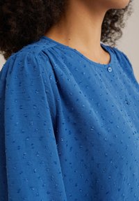 Blusa blu con texture, scollo rotondo, maniche corte e sottili motivi a punti in rilievo. Presenta una chiusura centrale con bottoni.