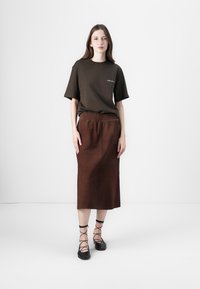 Bruine T-shirt met korte mouwen, relaxed fit, gecombineerd met een bruine midi-jurk met een elastische tailleband. Zwarte platte sandaaltjes maken de look compleet.