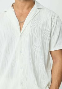 Chemise blanche à manches courtes arborant un motif ondulé texturé, un col en V, et des boutons transparents, avec une coupe décontractée et un tissu lisse.