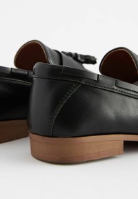 Mocassins en cuir noir avec une texture lisse, dotés d'un petit talon en caoutchouc brun et d'un gland décoratif à l'avant.