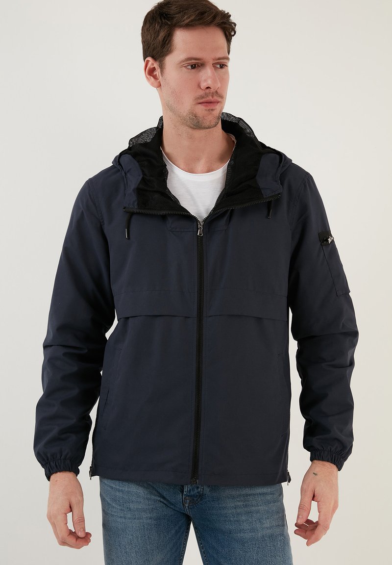 Temno moder hoodie z zadrgo spredaj, elastičnimi manšetami in mrežasto podlogo. Ima dva sprednja žepa in subtilen logotip na rokavu.