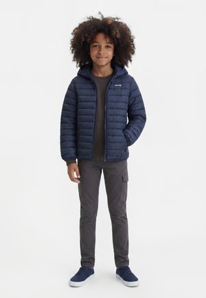 Barn med krøllet hår iført en mørkeblå hættetrøje med dunjakke, mørke cargobukser og navy sneakers, der står mod en ensfarvet baggrund.