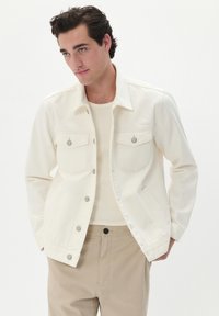 Jachetă din denim alb cu două buzunare la piept, accesorii din argint și guler clasic, asortată cu o cămașă bej cu nervuri și pantaloni bej deschis.