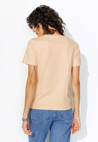 T-shirt en coton pêche à manches courtes, col rond, coupe décontractée, texture lisse, porté avec un jean en denim bleu, tatouages visibles sur les bras.