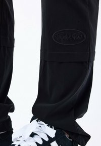 Pantaloni neri in un materiale leggero con vestibilità ampia, caratterizzati da un logo in rilievo sulla coscia sinistra e un dettaglio a polsino all'orlo.