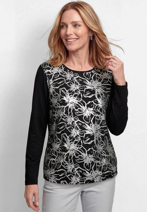 Frau mit blondem Haar, die ein schwarzes Langarmoberteil mit weißem Blumenmuster und hellgraue Hose trägt, lächelt, während sie ihr Haar berührt.