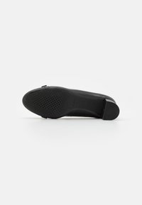 Semelle de chaussure en caoutchouc noir avec une finition lisse, présentant un motif de grip en points et un talon arrondi bas. Design minimaliste sans matériel visible.