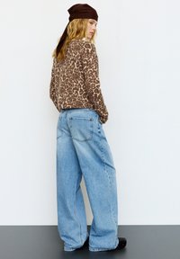 Brązowy sweter w panterkę, szerokie jasnoniebieskie dżinsy, czarne buty i brązowa chusta na głowie. Modelka stoi tyłem do kamery.