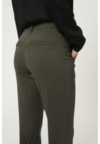 Personne portant un pantalon ajusté vert olive avec des poches passepoilées à l'arrière et une chemise noire à manches longues, main dans la poche latérale, sur un fond pâle.
