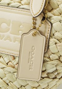 Beżowa skórzana metka z wytłoczonym napisem „COACH NEW YORK”, przymocowana złotym łańcuszkiem do tkanej słomkowej tła.