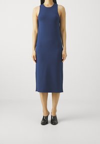 Kobieta stoi w bezrękawowej, granatowej midi sukience z czarnymi butami na obcasie, na tle białego tła.