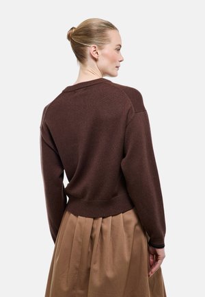 Femme aux cheveux blonds attachés en chignon, portant un pull en laine marron foncé et une jupe plissée camel taille haute, vue de dos avec la tête tournée vers la droite.
